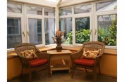 Two wicker chairs with red cushions flank a small table by glass-enclosed windows.