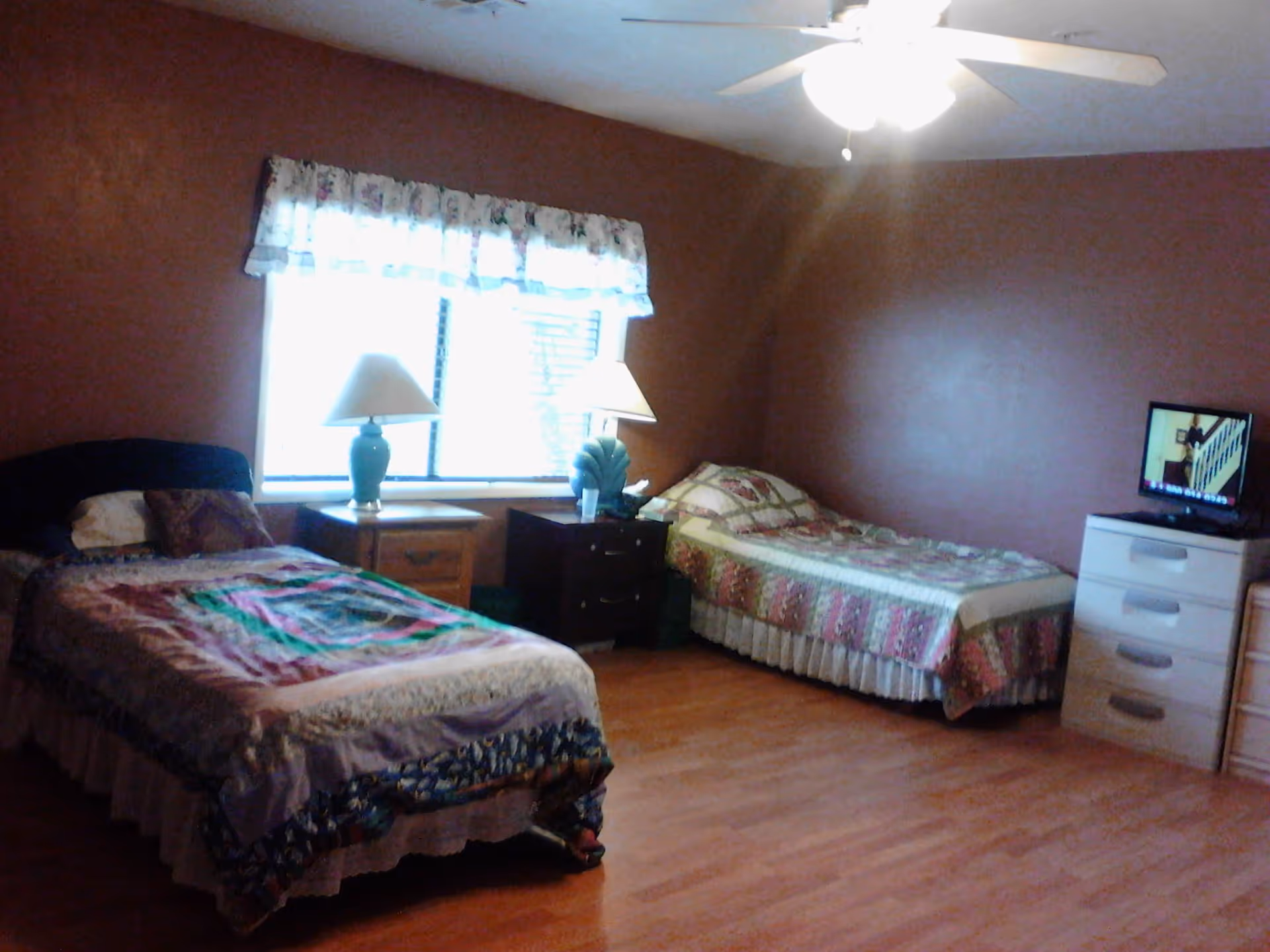 A bedroom with two single beds covered with patterned quilts, a window with floral curtains, two bedside tables each with a lamp, a ceiling fan with light, and a small dresser with a television on top.