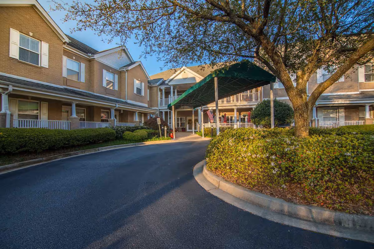 Exterior view of The Gardens at Eastside senior living facility showing a two-story building with beige brick walls, white window shutters, a covered entrance with a green canopy, and landscaped bushes and trees along a curved driveway.