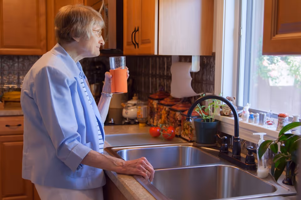 Elderly woman standing at a kitchen sink holding a glass and looking out the window.