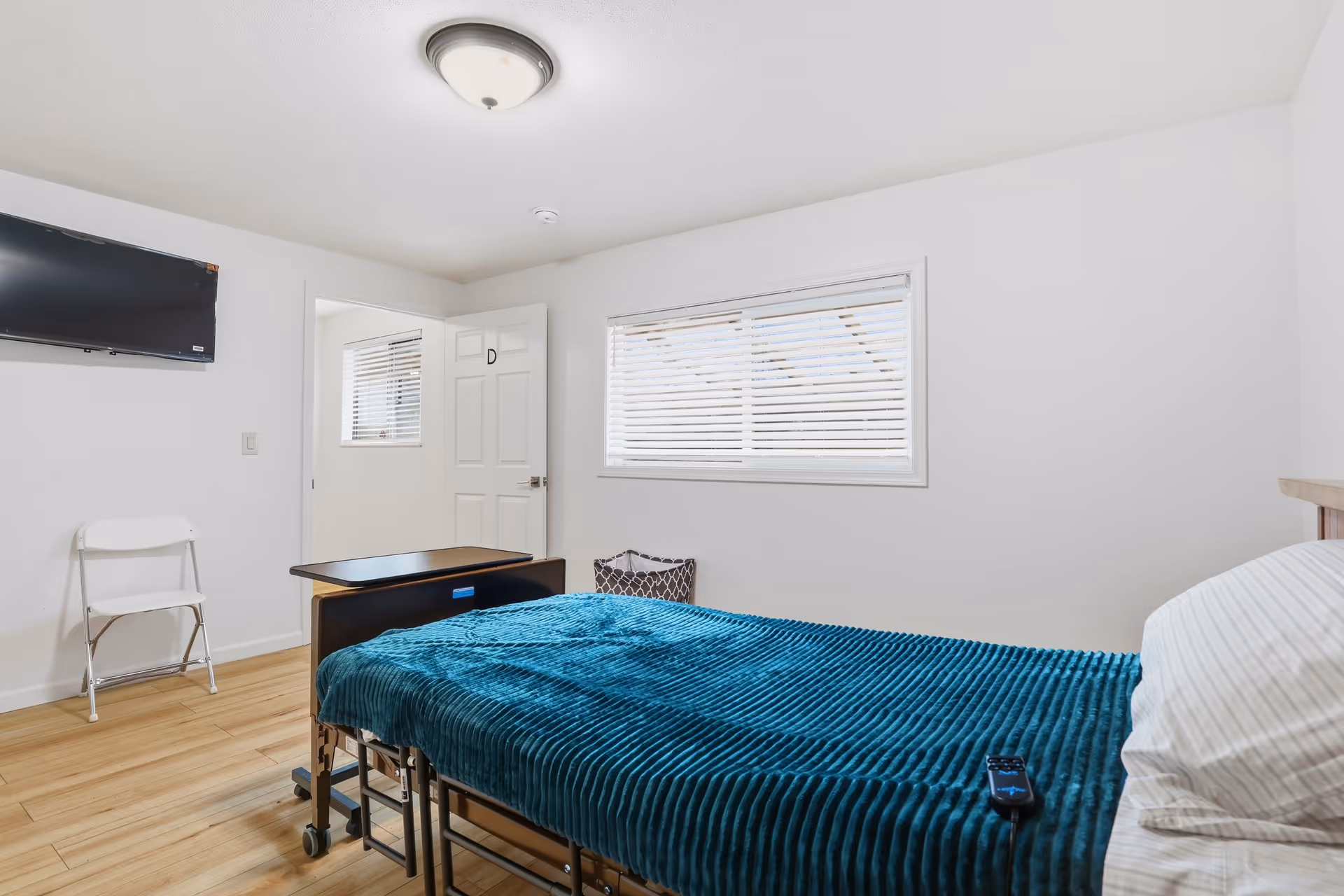 A simple bedroom with a bed, teal cover, window blinds, wall TV, and a small chair.