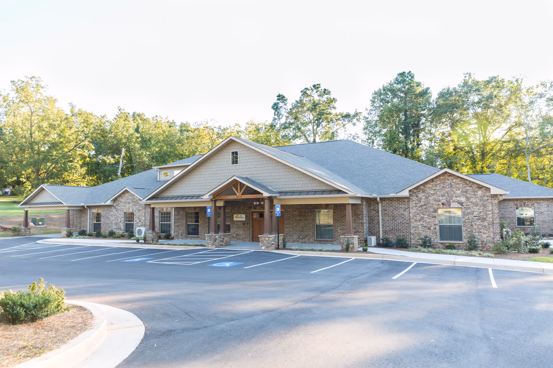 Exterior view of a single-story brick and stone building with a gabled roof, surrounded by trees and an empty parking lot with marked handicap spaces in front.