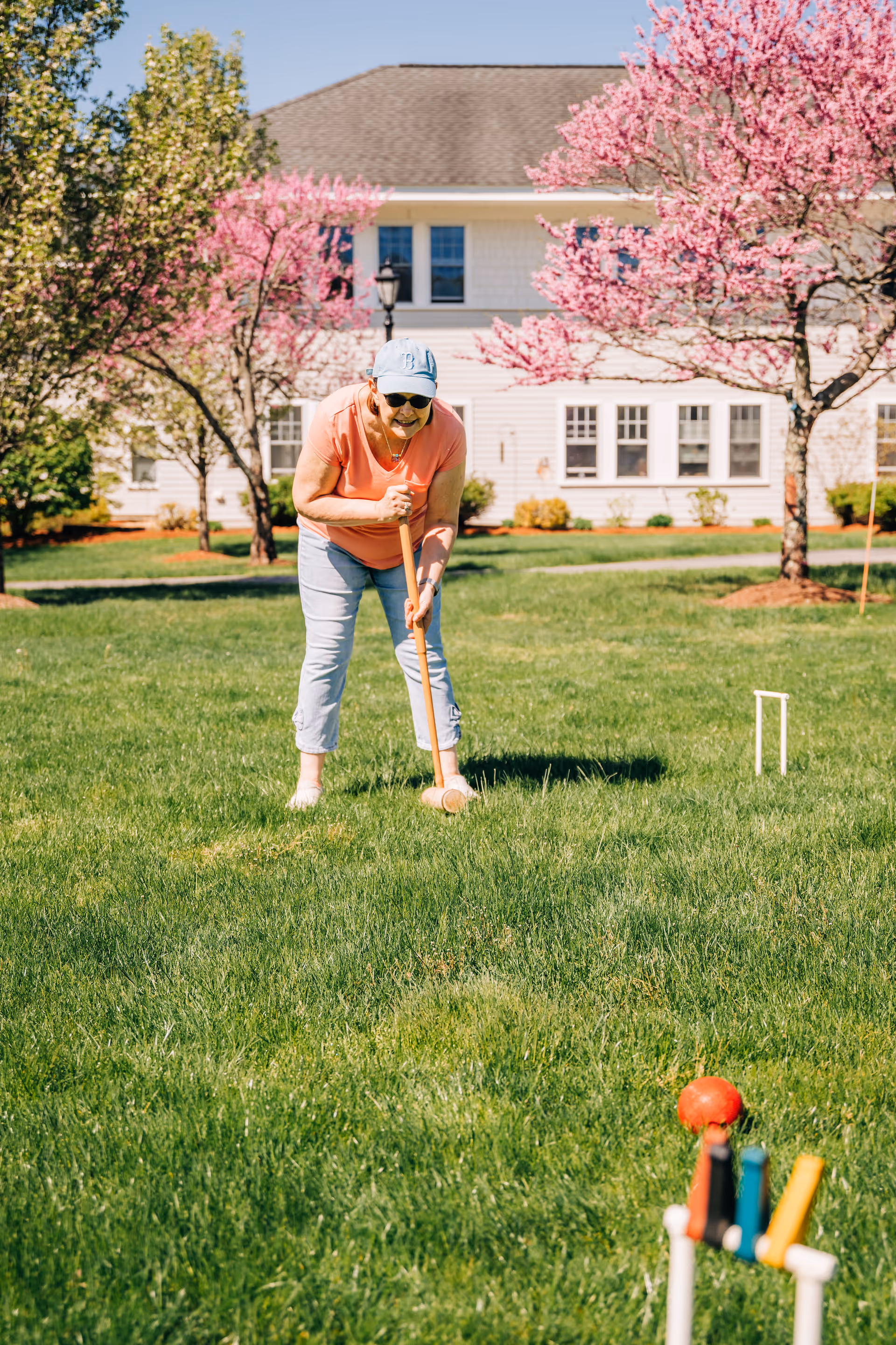 Senior woman plays croquet on a sunny lawn in front of a residence.