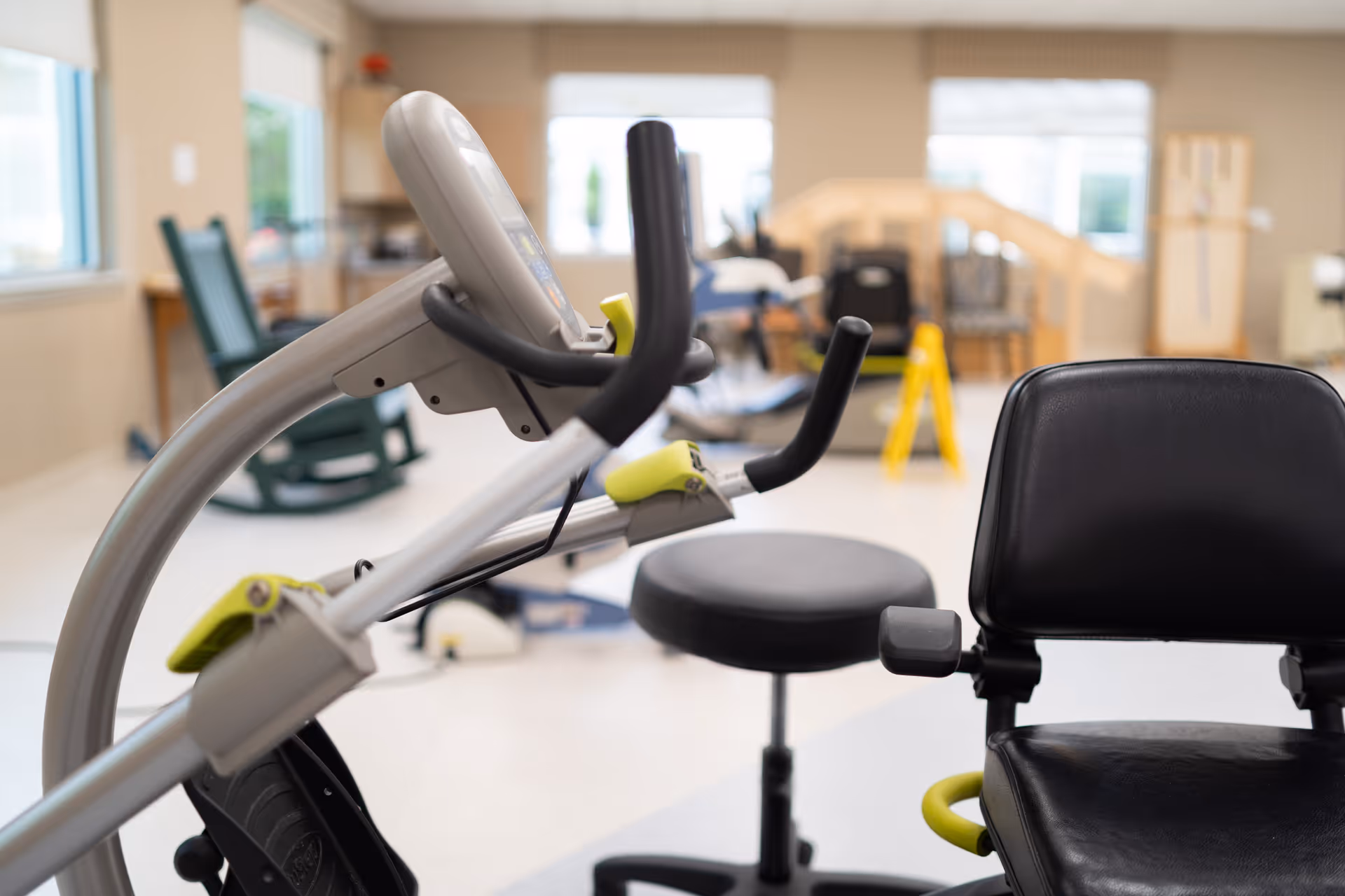 Indoor exercise room with stationary exercise bikes, a black padded chair, and a stool. The room has large windows letting in natural light and some chairs and equipment in the background.