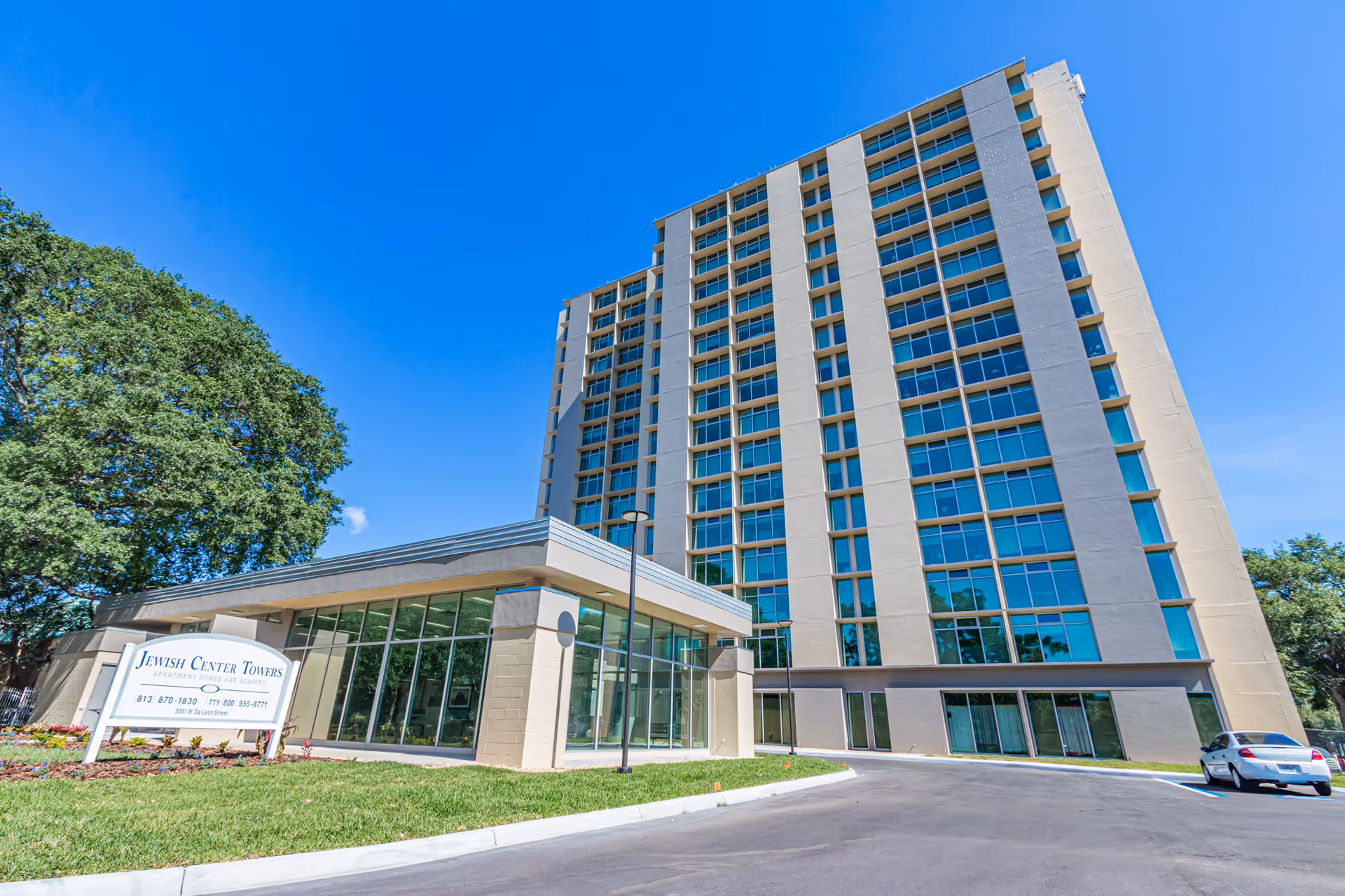 Exterior view of Jewish Center Towers, a tall multi-story senior apartment building with large windows, a smaller glass-walled entrance area, a sign with contact information, a tree on the left, and a car parked on the right under a clear blue sky.