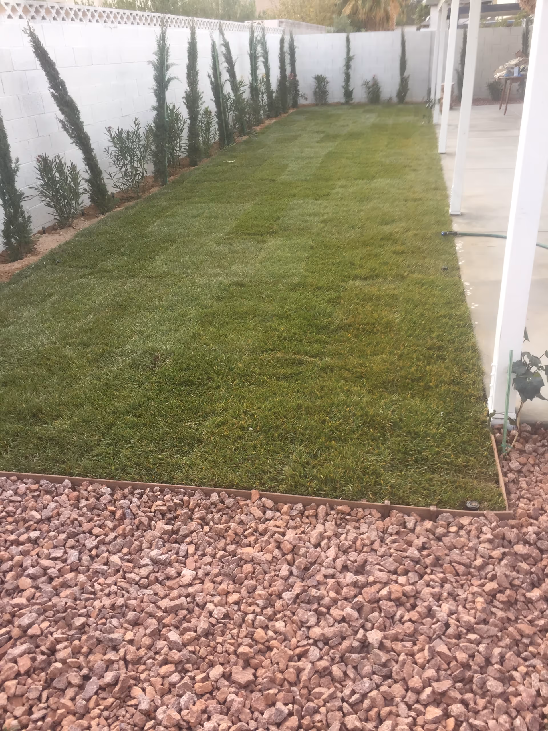 Narrow backyard with green lawn, gravel edges, white fence, and a covered patio area.