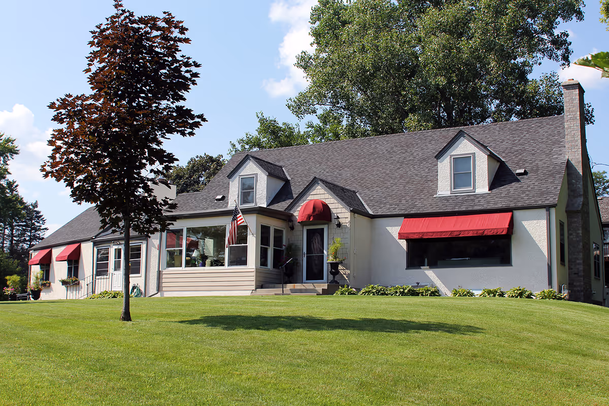 Exterior view of a single-story residential building with a gray roof, white walls, and red awnings over the windows. There is a well-maintained green lawn in front and a tree on the left side. The building has a front entrance with a small set of stairs and an American flag displayed near the door.