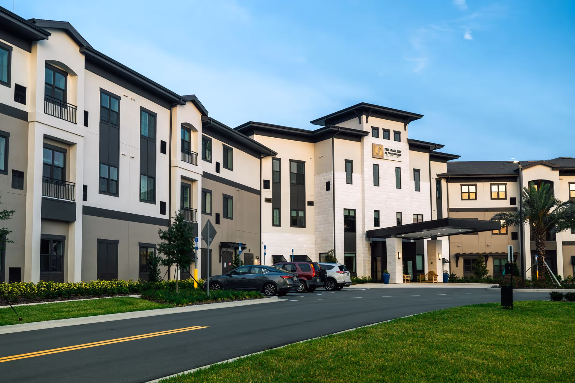 Front view of The Gallery at Port Orange, a three-story cream-and-gray senior living building with a covered entrance, parked cars, and landscaping under a blue sky.