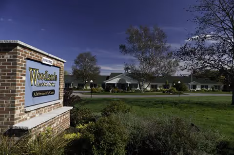 Brick sign reading Woodlands Memory Care in front of landscaped grounds with the facility building and trees behind it under a blue sky.