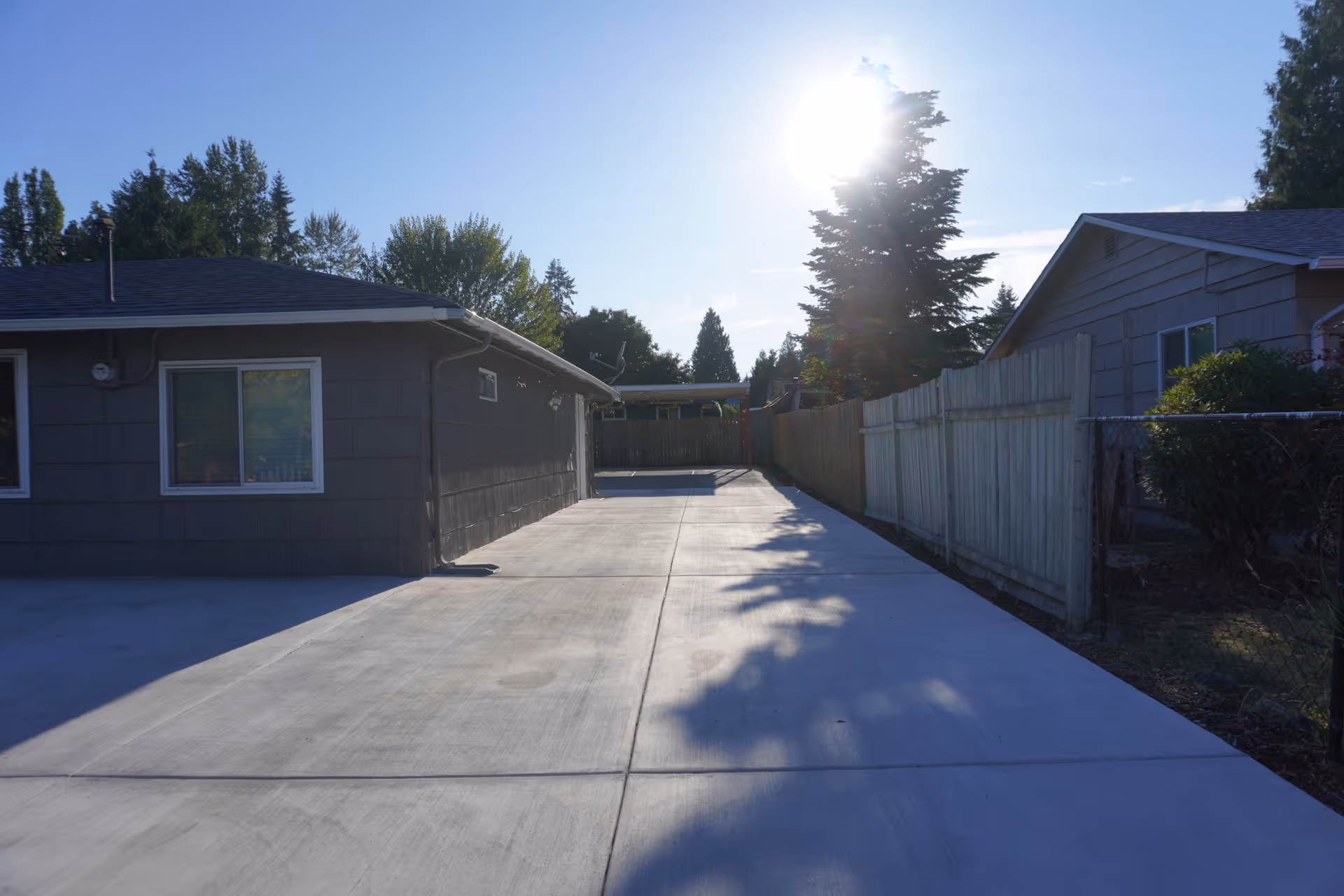 Wide concrete driveway runs between single-story buildings, with fences and trees in bright sunlight.