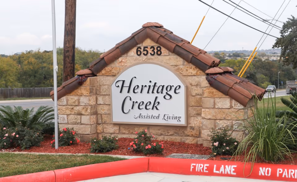 Stone entrance sign reading "Heritage Creek Assisted Living" with address number 6538, landscaping, and a red curb marked "FIRE LANE".