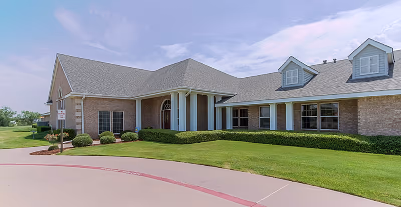 Exterior front view of a single-story brick building with a gray shingled roof, white columns at the entrance, multiple windows, and well-maintained green lawn and shrubs under a partly cloudy sky.