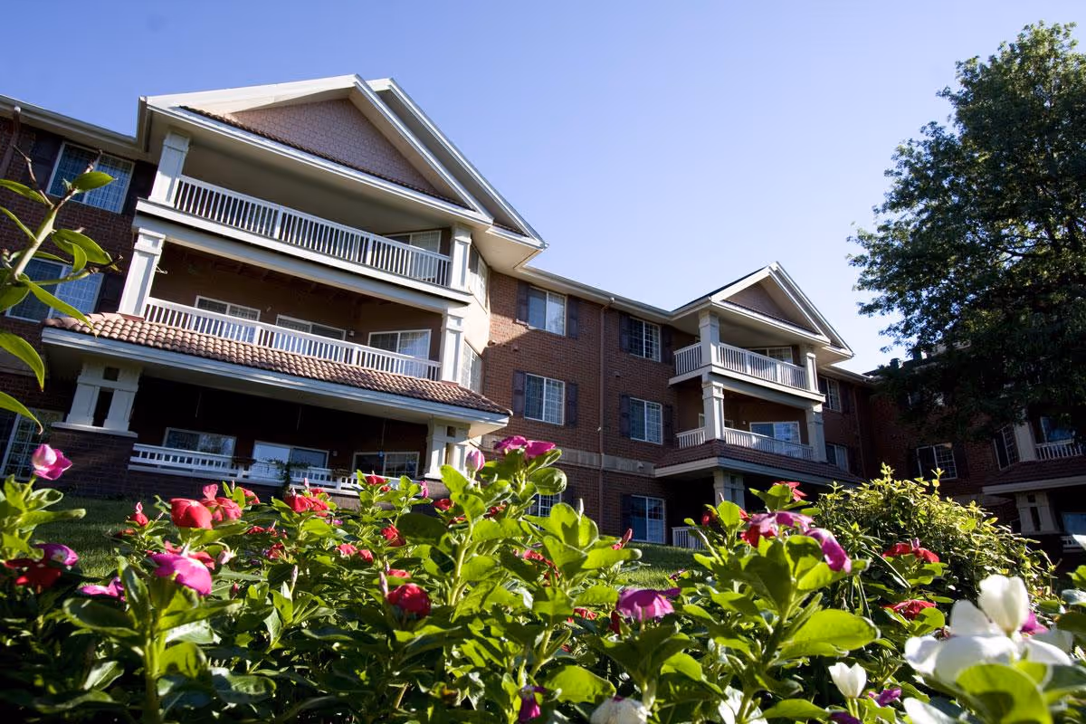 Exterior view of a multi-story brick residential building with balconies and flowering shrubs in the foreground under a clear sky.