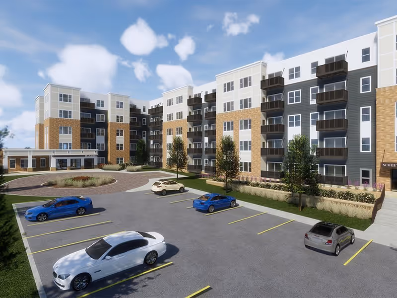 Exterior view of a modern multi-story senior living facility with a parking lot in front containing several parked cars. The building features a combination of brick and light-colored siding with multiple balconies and large windows under a partly cloudy sky.