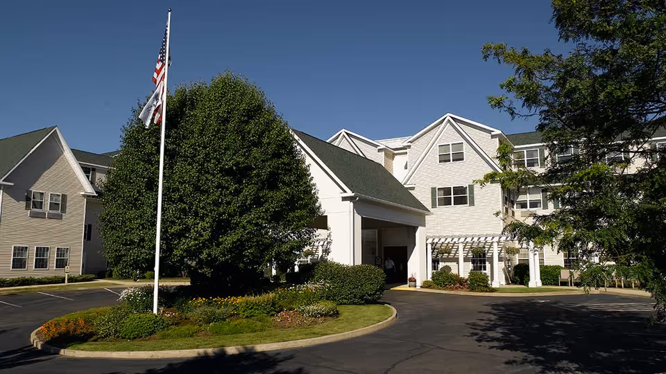 Front entrance of a multi-story senior living building with a flagpole, circular driveway, and landscaped greenery.
