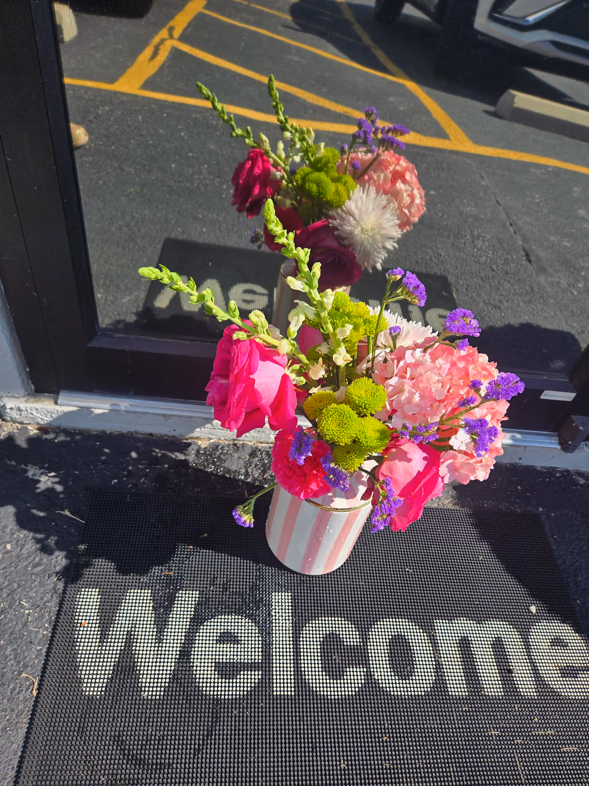 Colorful flower arrangement in striped vase on a welcome mat outside a glass door.