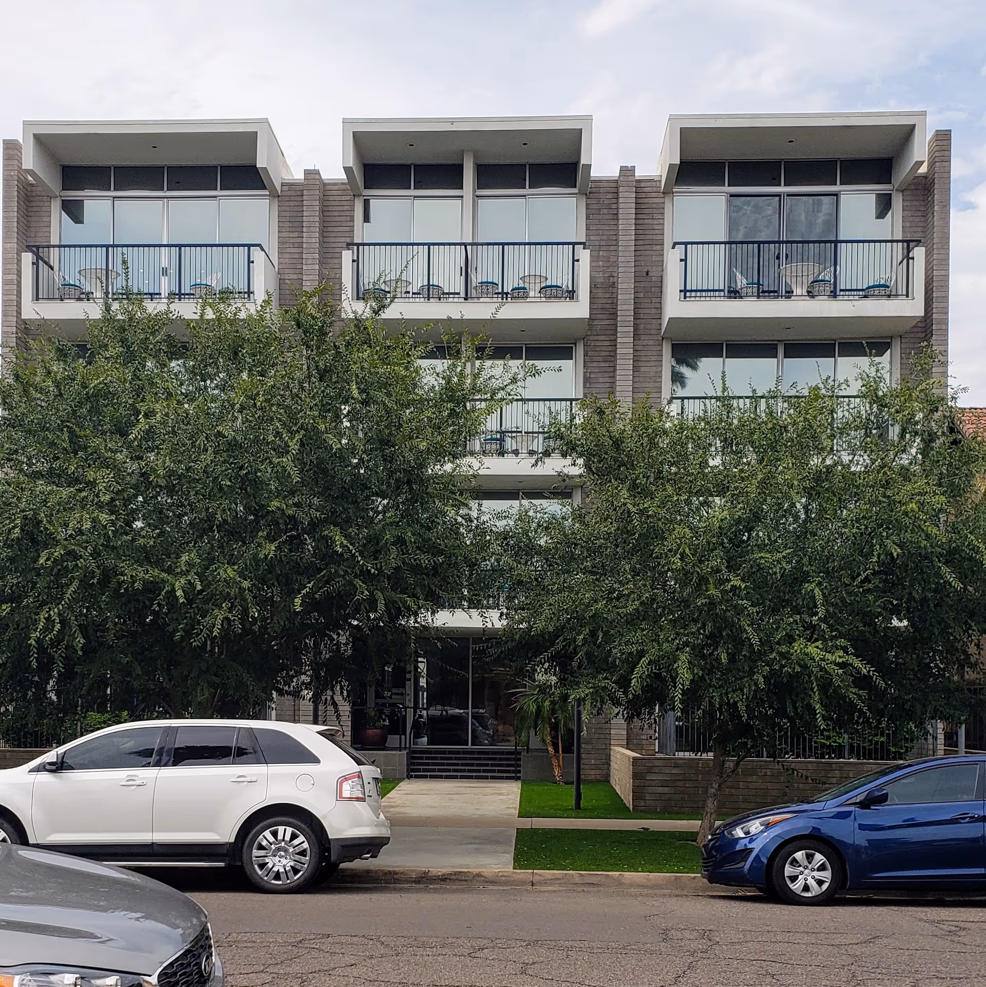 Modern senior living building with balconies, trees, and parked cars along the street.