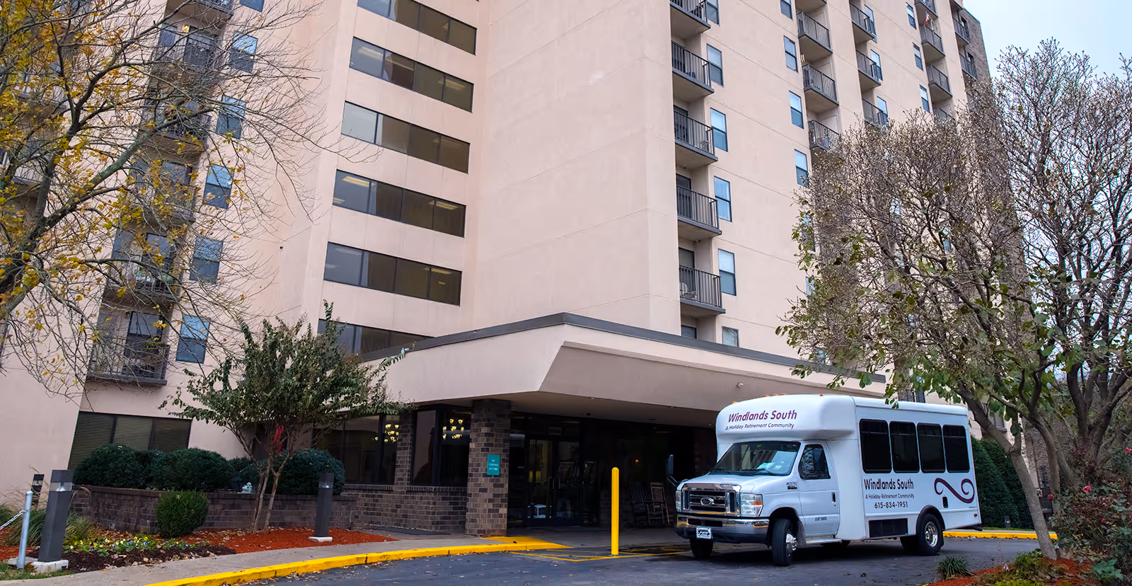 Exterior view of Windlands South, a multi-story senior living facility with a covered entrance. A white shuttle bus with Windlands South branding is parked in front. Trees with sparse leaves and landscaped bushes surround the entrance area.