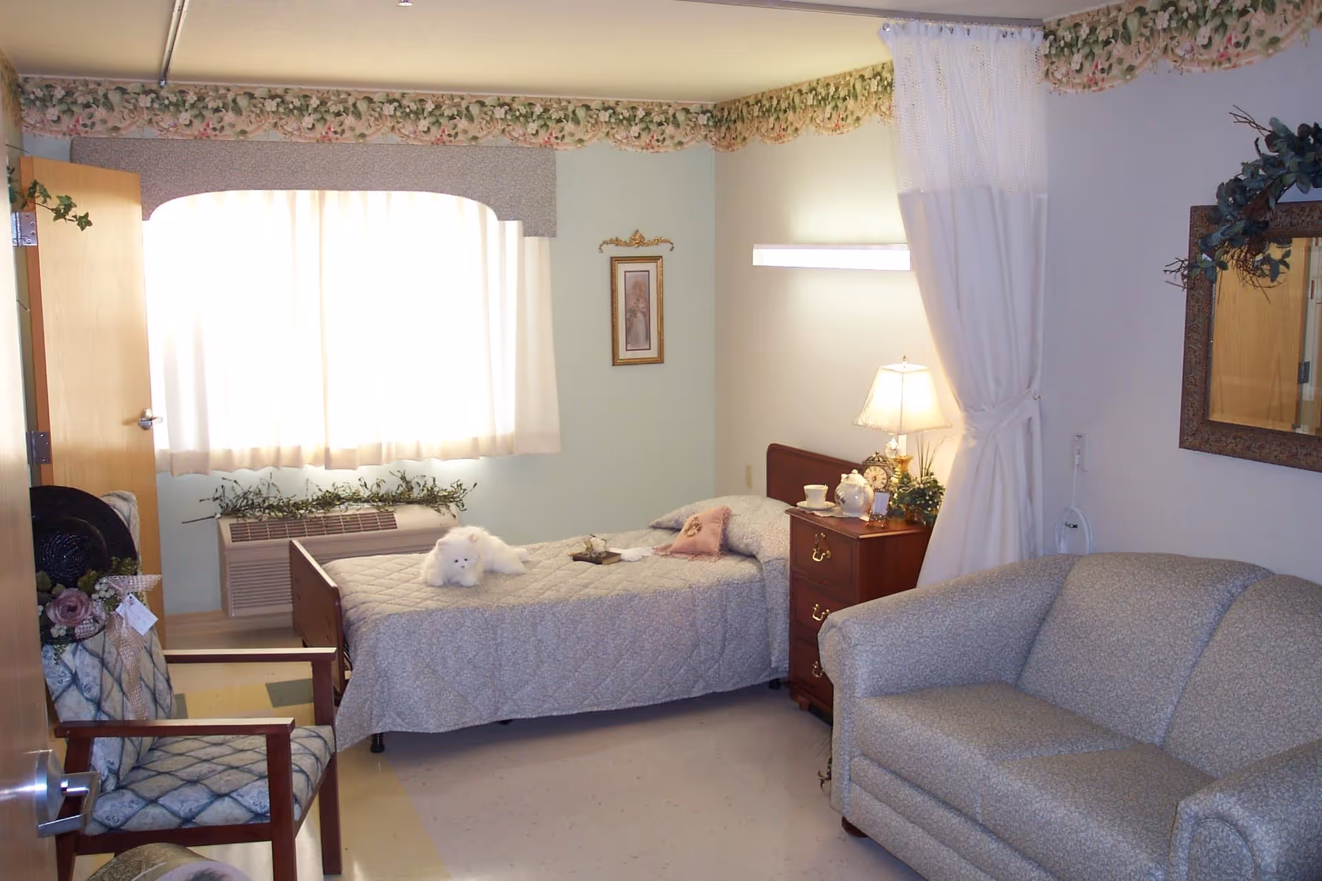 A cozy senior living bedroom with a single bed, nightstand and lamp, armchair and sofa beneath a curtained window.