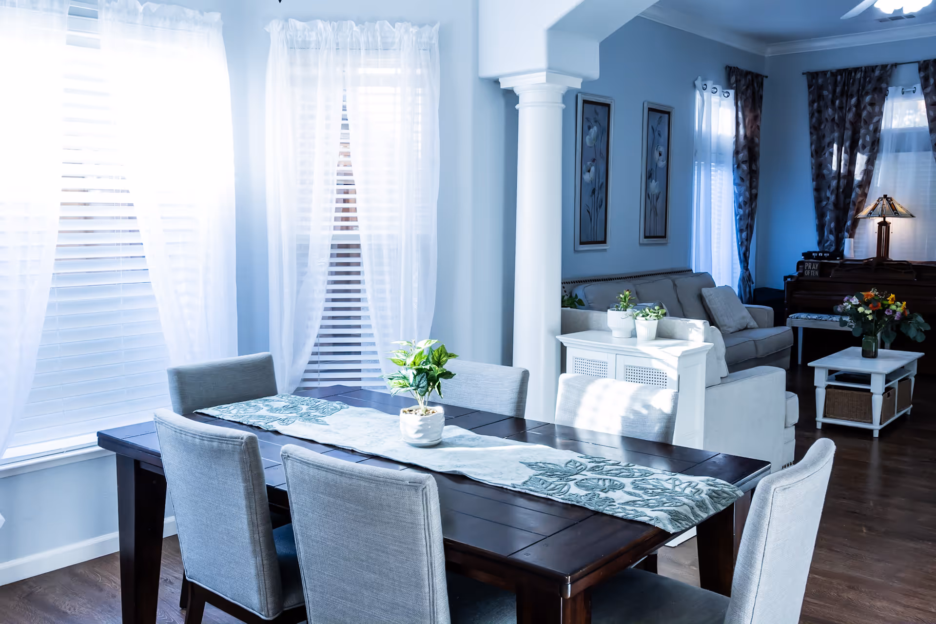 A bright dining area with a dark wooden table and six gray upholstered chairs. The table has a decorative runner and a small potted plant. Large windows with sheer white curtains allow natural light to fill the room. In the background, there is a living room area with a white sofa, a white side table with potted plants, framed floral artwork on the wall, and a piano with a lamp and a vase of flowers.