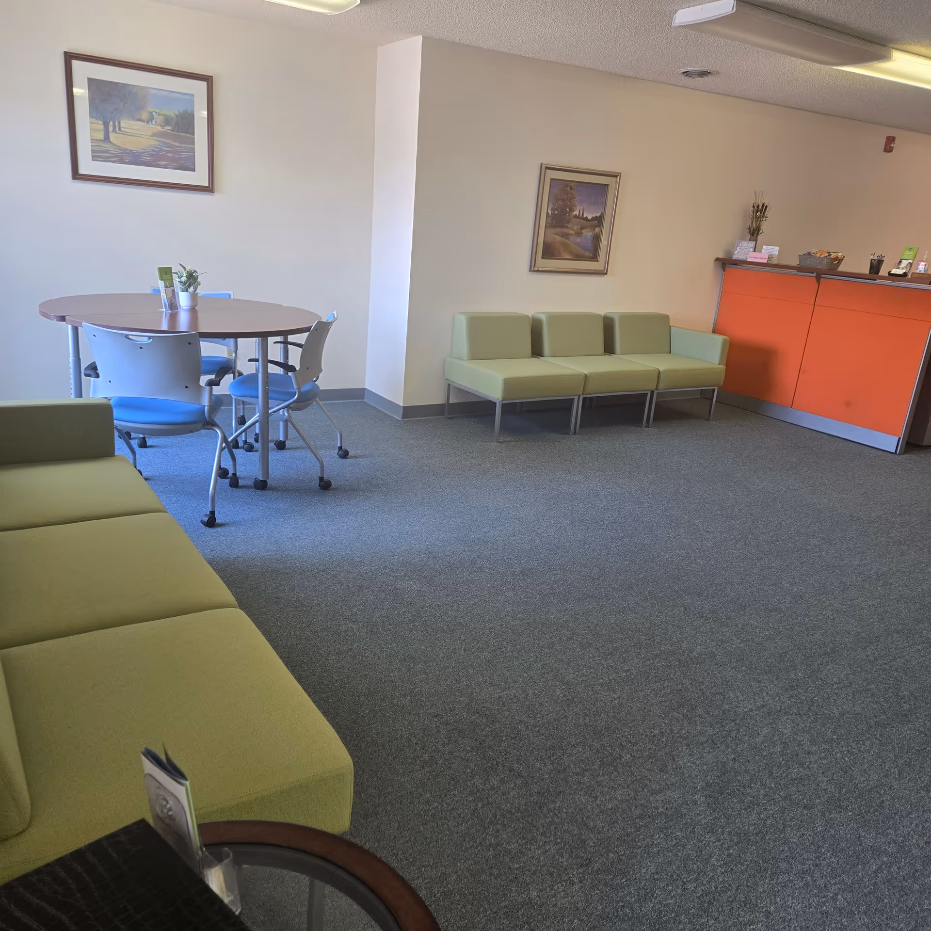 Communal lounge in a senior living facility with lime-green couches, a round table, and blue chairs.