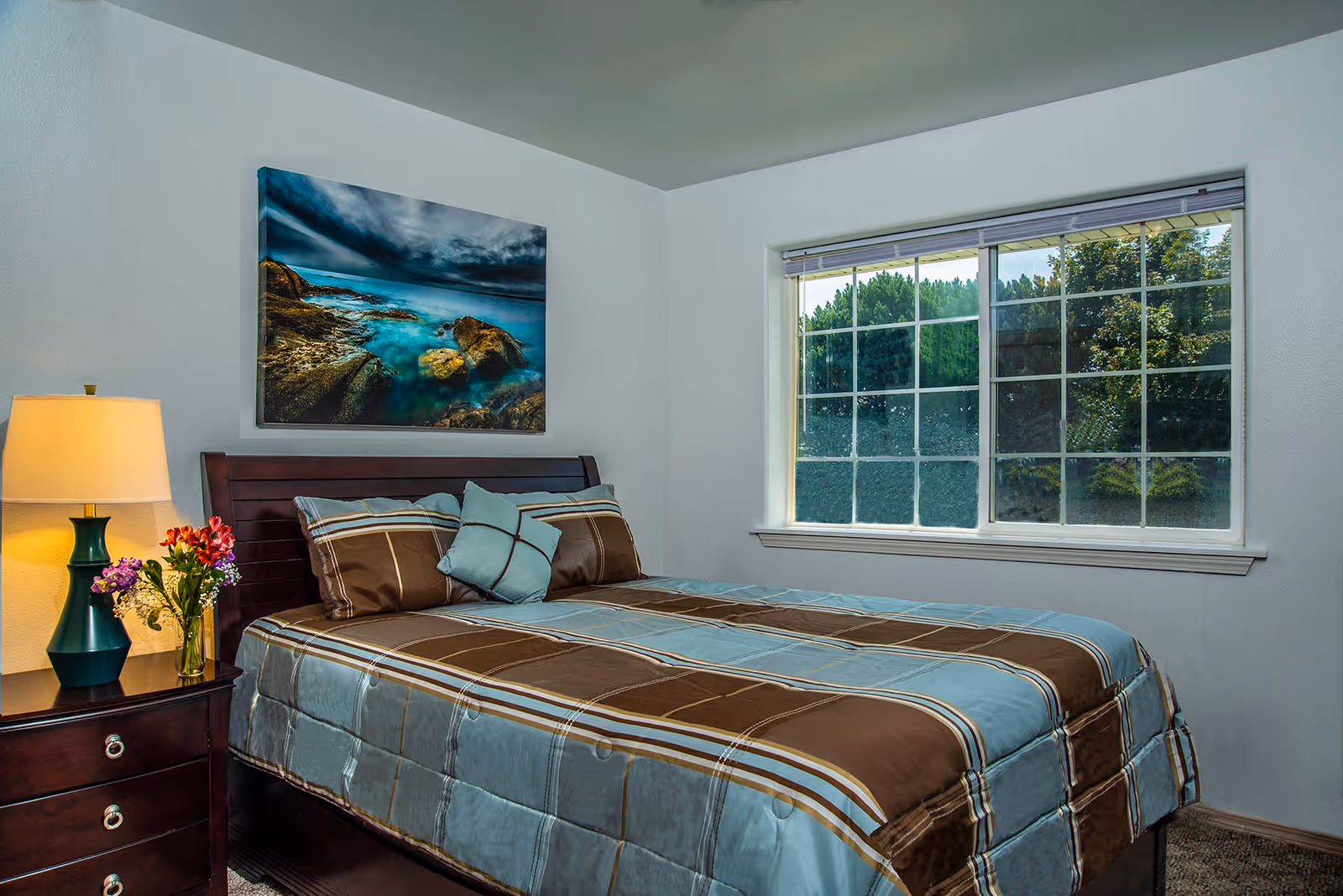 A neatly made bed with a brown and blue striped comforter and matching pillows in a bright bedroom. There is a wooden nightstand with a green lamp and a vase of colorful flowers next to the bed. A large window with white trim lets in natural light, and a framed picture of a rocky coastline hangs on the wall above the bed.