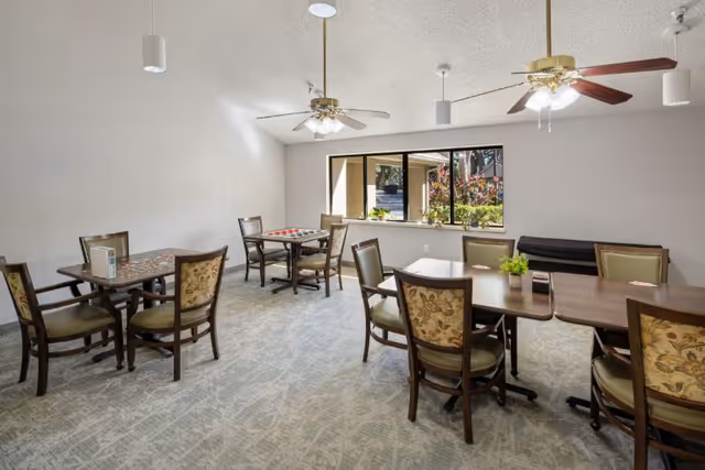 Interior dining room with several tables, chairs, and ceiling fans in a senior living facility.