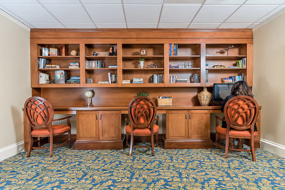 A study or computer area with a large wooden built-in bookshelf and desk. The bookshelf contains various books and decorative items. Three wooden chairs with orange cushions are placed in front of the desk, and a person is seated at the rightmost chair using a computer. The room has a patterned blue and gold carpet and beige walls.