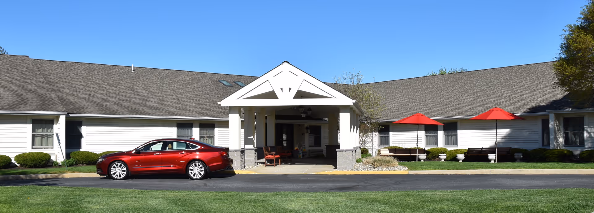 Front exterior view of a single-story senior living facility building with a covered entrance, a red car parked in front, and two red umbrellas shading benches on the right side. The building has white siding and a gray shingled roof under a clear blue sky.