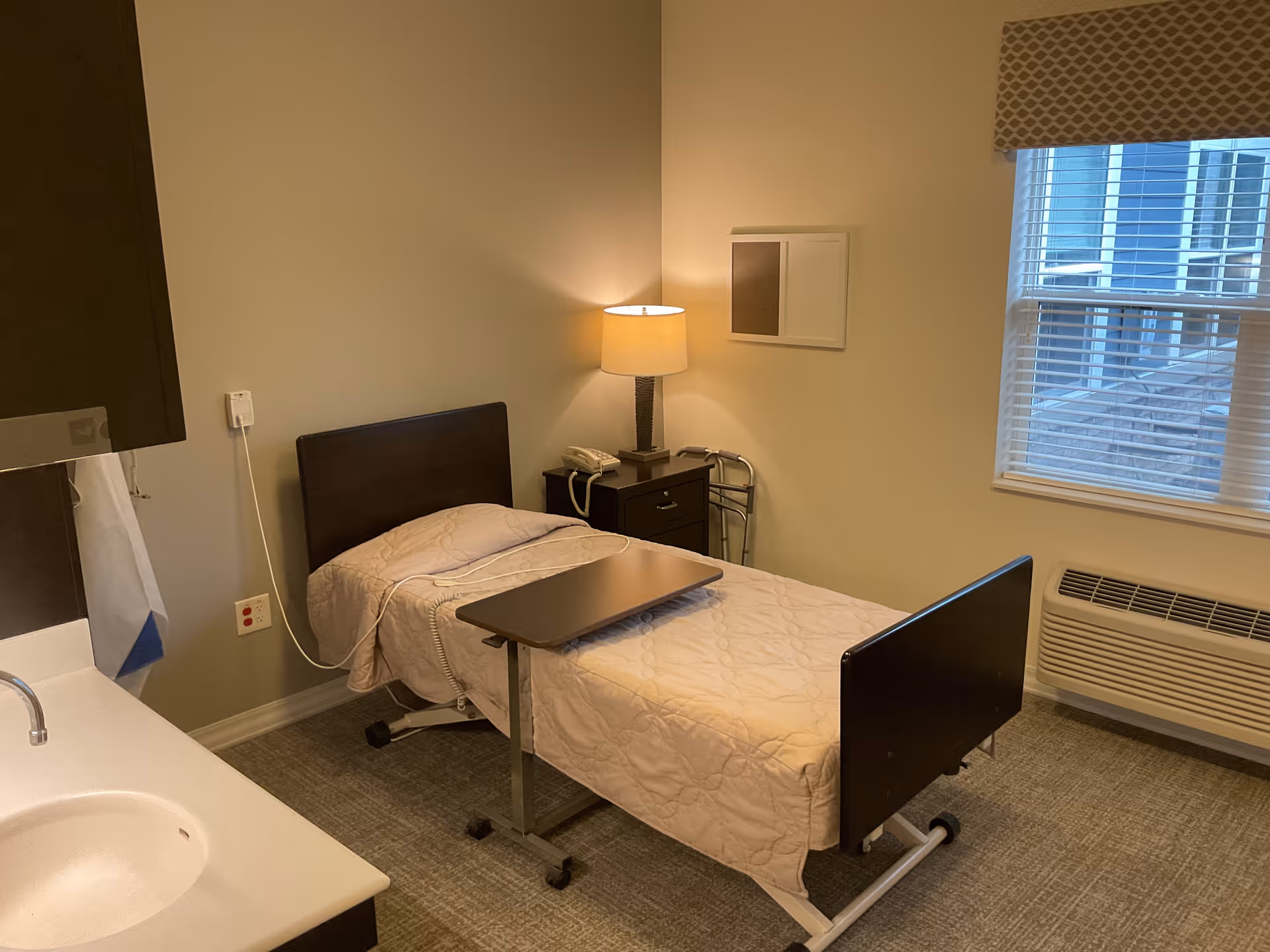 A single patient bedroom with a bed, nightstand, lamp, window, and sink area.