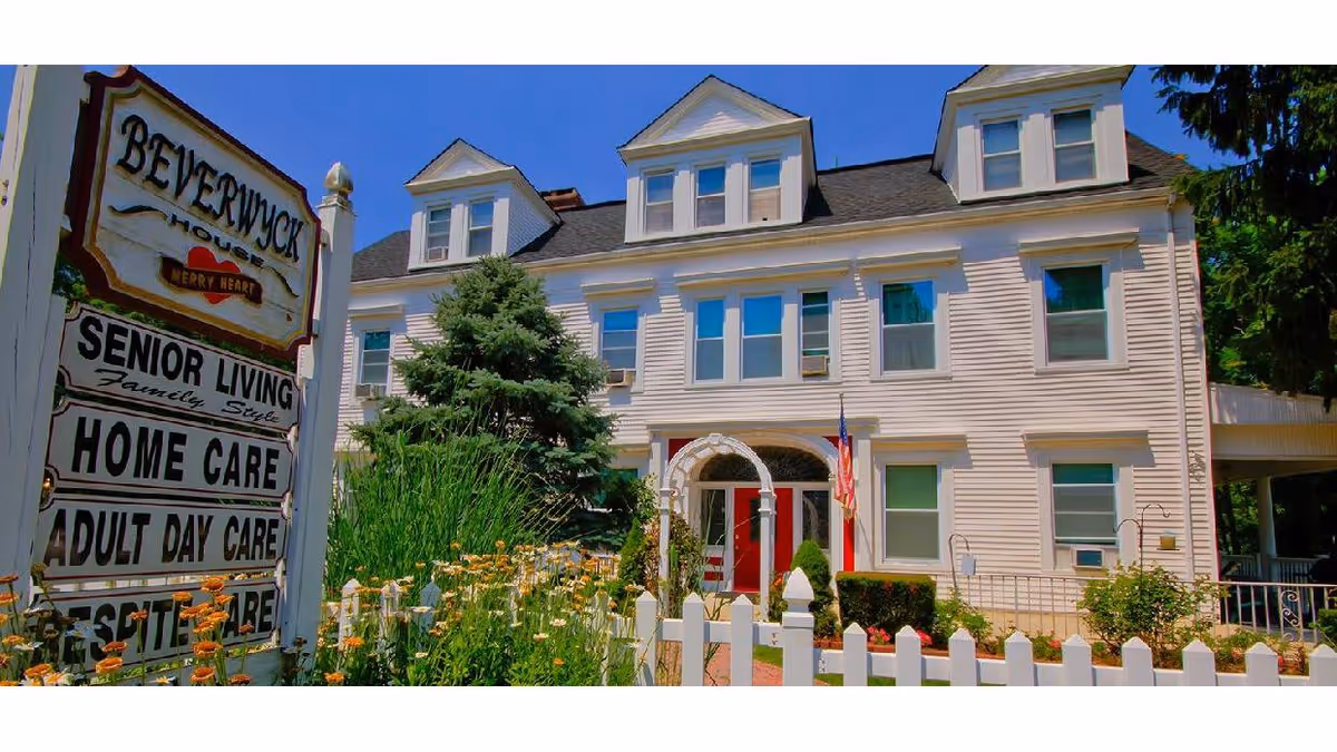Exterior view of a large white senior living facility building with multiple windows and dormers on the roof. There is a white picket fence and a garden with flowers in front. A sign near the entrance reads 'Beverwyck House of Merry Heart' and lists services including senior living, home care, adult day care, and respite care. An American flag is displayed near the entrance.