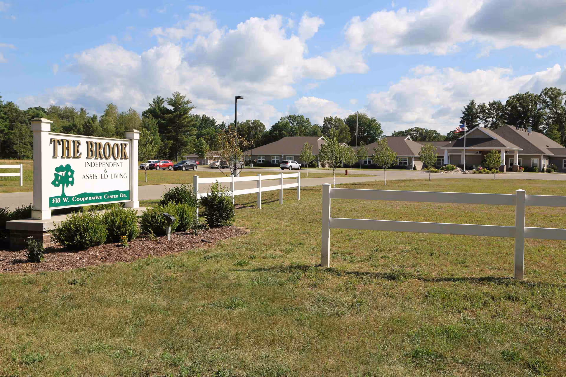 Exterior view of The Brook of Newaygo senior living facility showing a large sign with the facility name and address, a white fence, a grassy area, a parking lot with cars, and a single-story building under a partly cloudy sky.