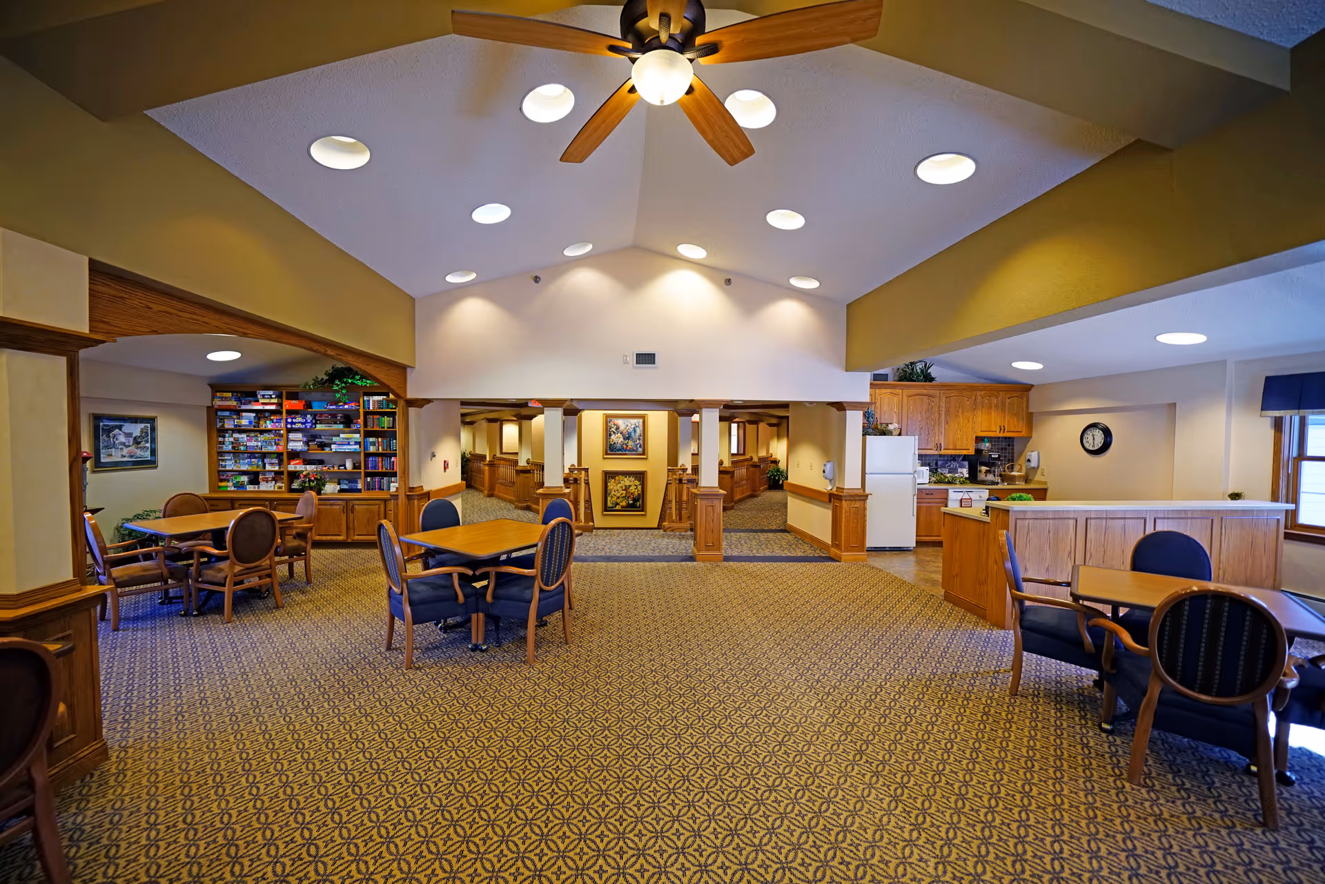 Open, well-lit senior living lounge with round tables, chairs, and a kitchenette.