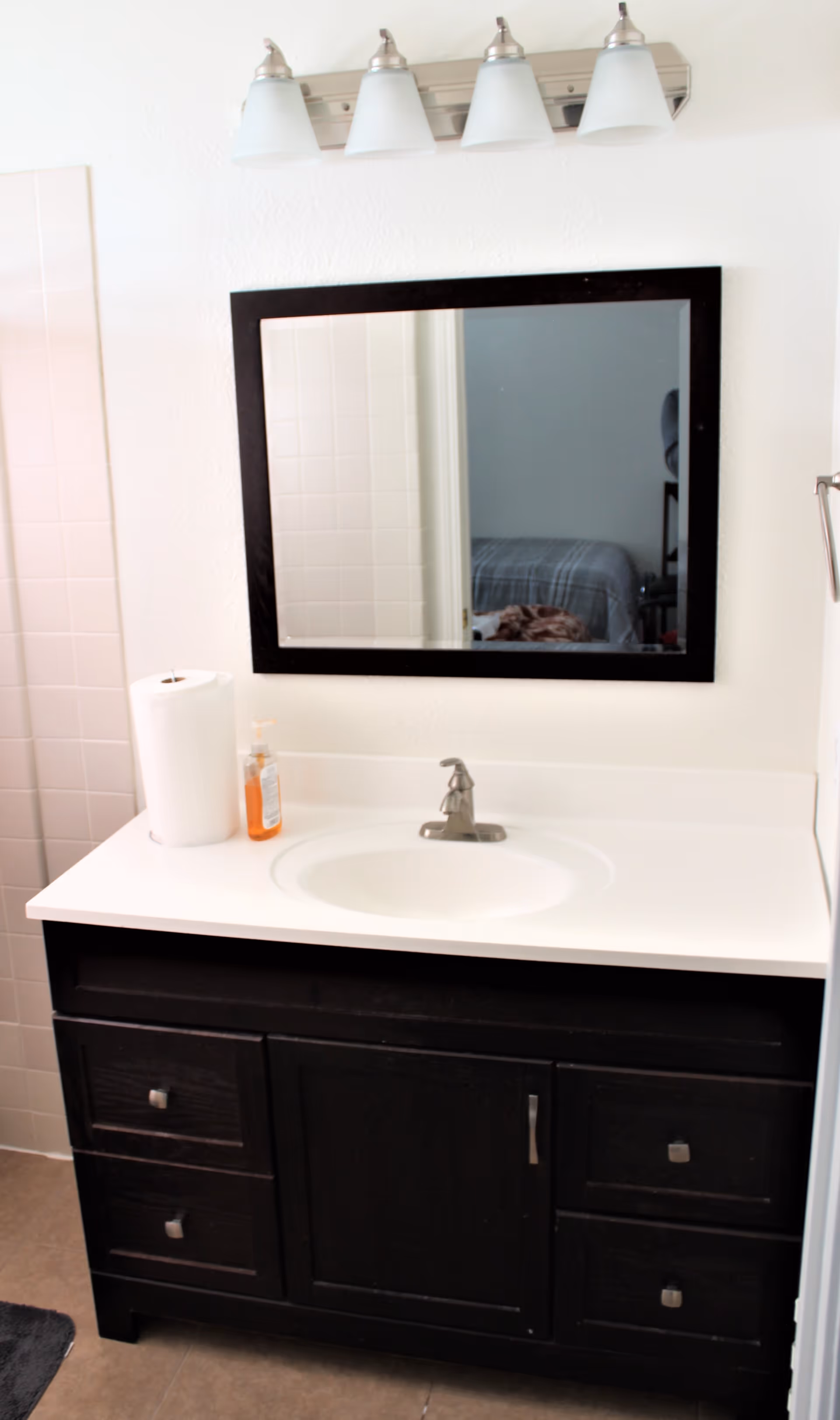 Bathroom vanity with a white countertop, a built-in sink, and a silver faucet. Above the vanity is a rectangular mirror with a black frame and a four-light fixture with frosted glass shades. To the left of the vanity is a roll of paper towels and a bottle of orange liquid soap. The reflection in the mirror shows part of a bedroom with a bed covered in a gray blanket.