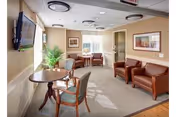 A cozy senior living lounge with beige walls, seating, a wall-mounted TV, and plants.