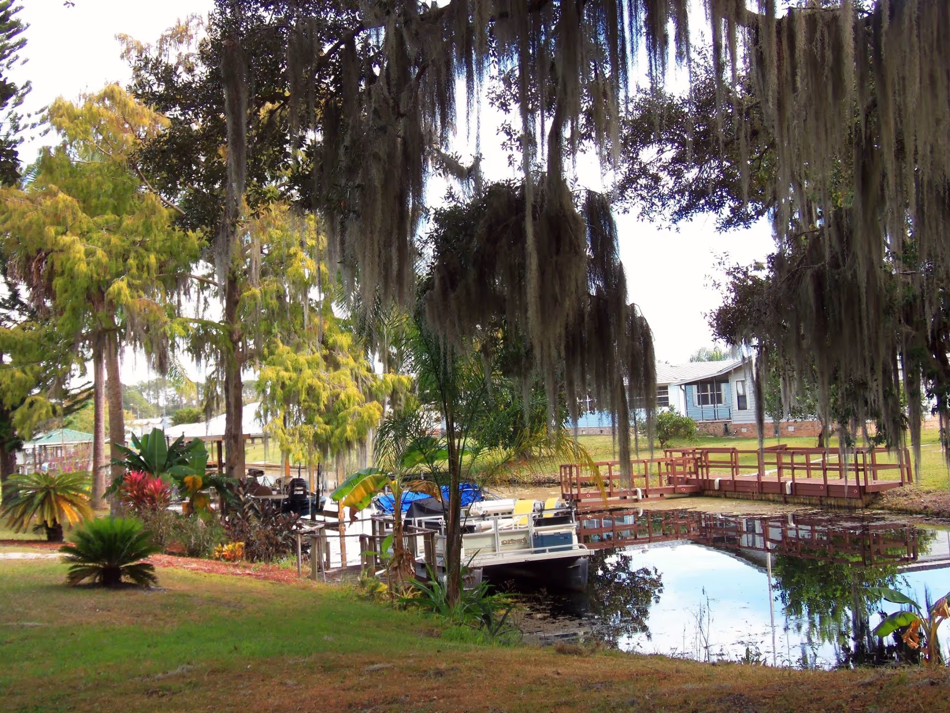 A peaceful outdoor scene at Palms Estates of Highlands County featuring a small dock with a boat on a calm pond surrounded by lush greenery, trees draped with Spanish moss, and a light blue building in the background.