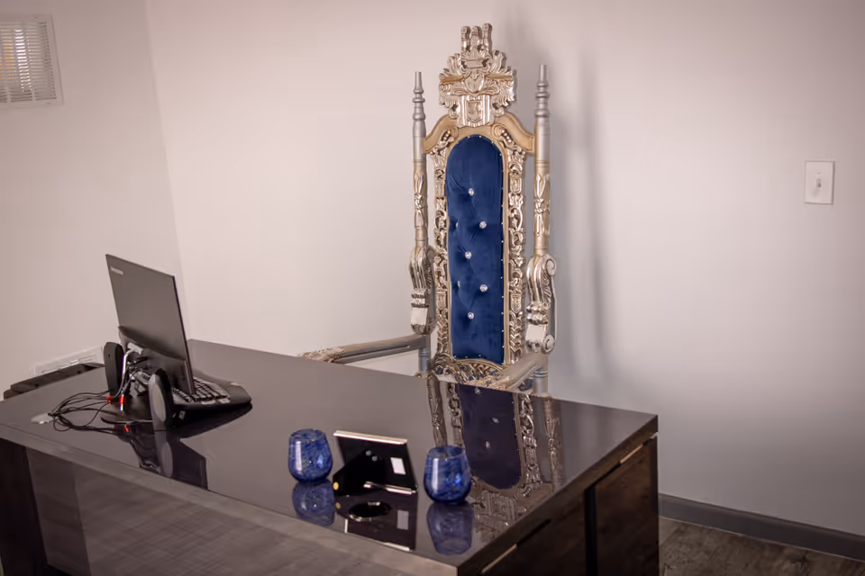 Ornate silver throne chair behind a dark desk with computer in a plain office.