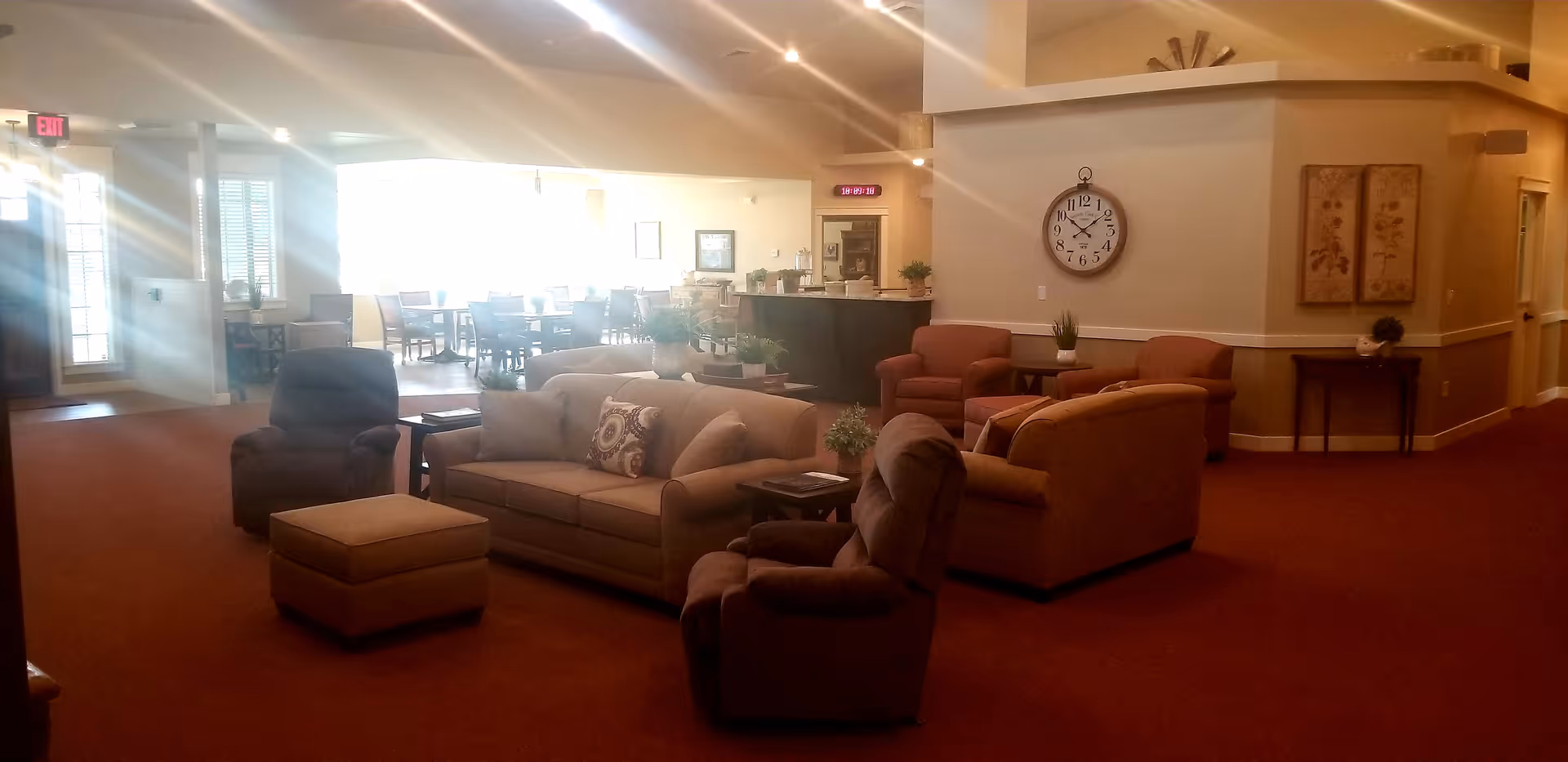 A spacious senior living facility common area with comfortable seating including sofas and armchairs arranged around small tables. The room has warm lighting, a large wall clock, decorative wall art, and a dining area with tables and chairs visible in the background near large windows letting in natural light.