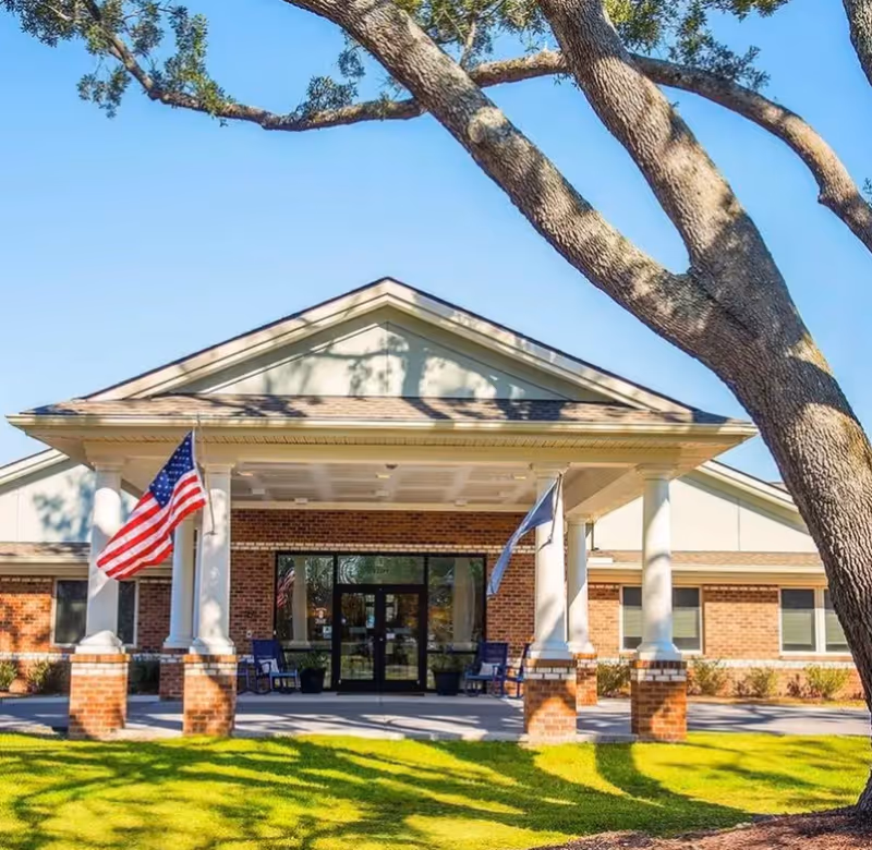 Front exterior view of Inlet Oaks Assisted Living of Murrells Inlet building with a covered entrance supported by white columns, two flags on either side, and a large tree in the foreground.