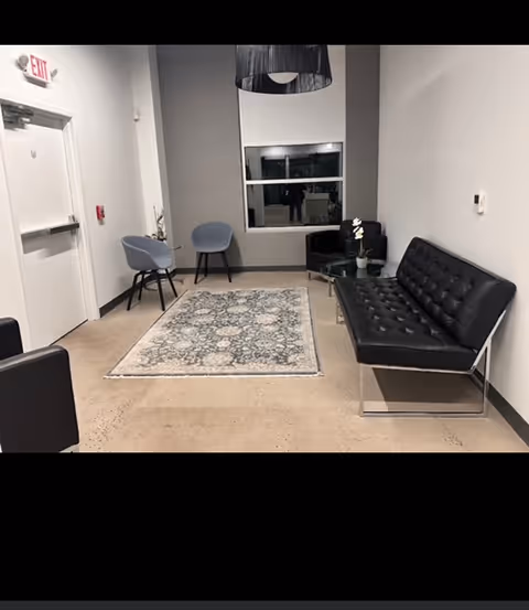 Modern waiting area with black leather sofa, blue chairs, rug, and windowed wall.
