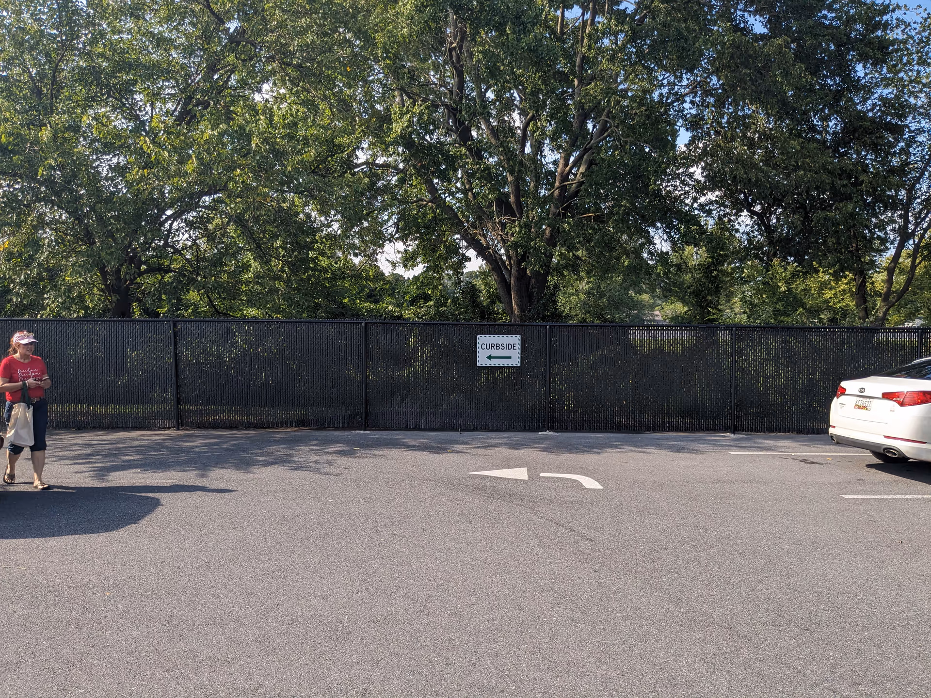 Outdoor parking area with a tall black fence, trees, a curbside sign, and a parked car.