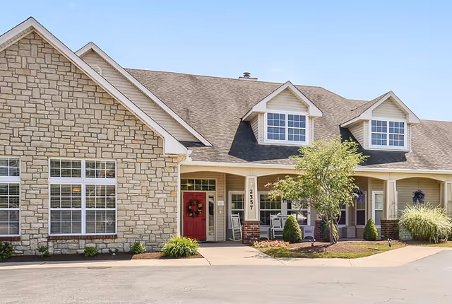 Front exterior of a stone-faced senior living building with a red entrance door, covered porch with chairs, dormer windows, and small landscaped beds.