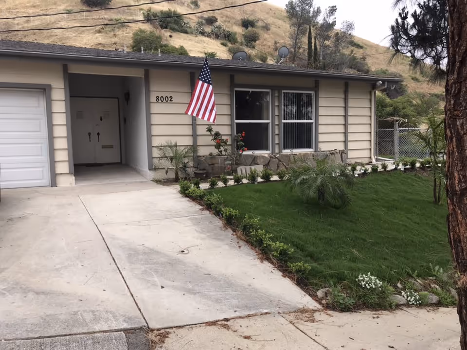 Front exterior of a single-story house with an American flag, driveway, garage, and a grassy front yard by house number 8002.
