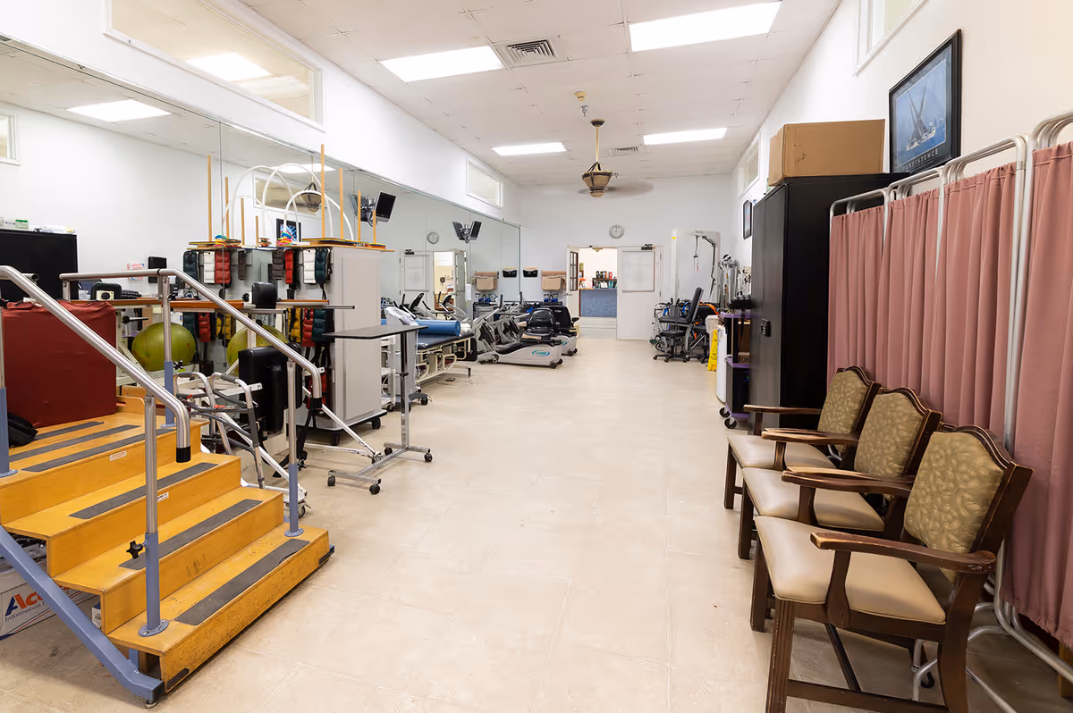 Spacious interior therapy room with exercise equipment, parallel bars, treatment tables, and a row of chairs by privacy curtains.