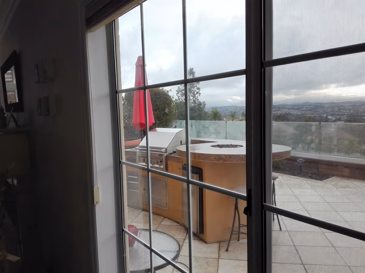 View through a glass sliding door onto a patio with an outdoor grill and round countertop, a red umbrella, and a distant cityscape.