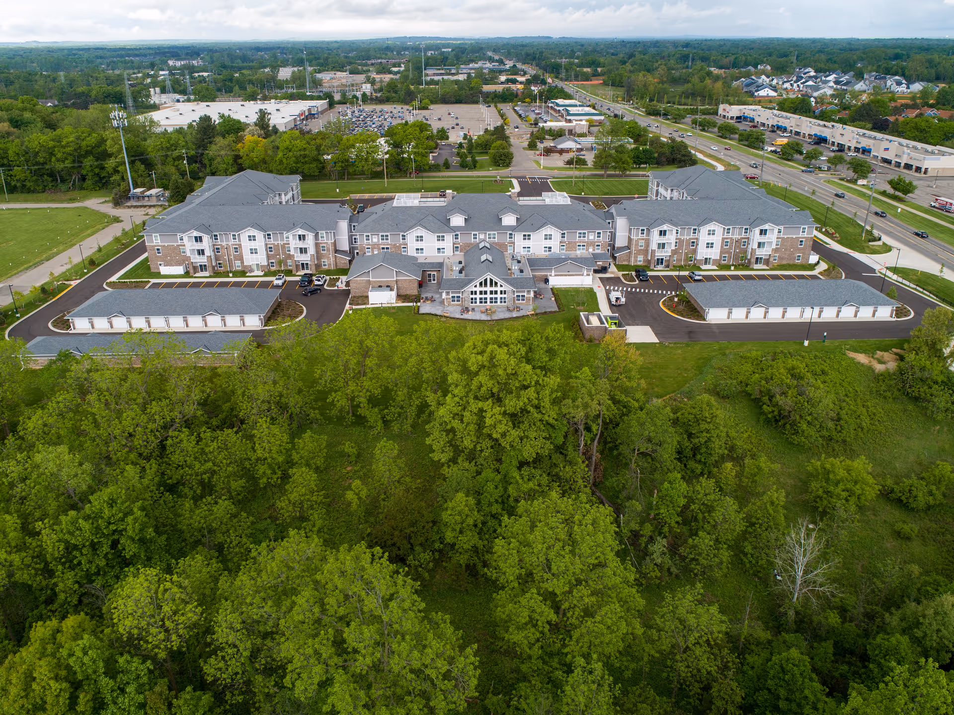 Aerial view of a three-story retirement community building with parking lots, twin garages, surrounding trees, and nearby roads.