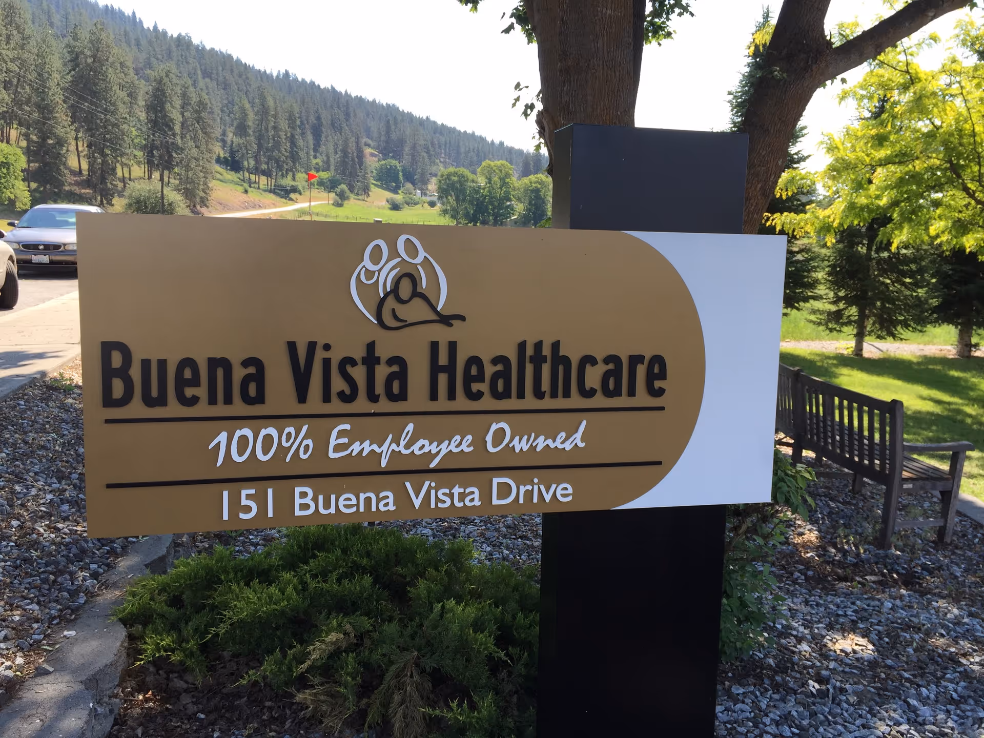 Outdoor sign reading "Buena Vista Healthcare — 100% Employee Owned — 151 Buena Vista Drive" with a bench, trees, and hills in the background.