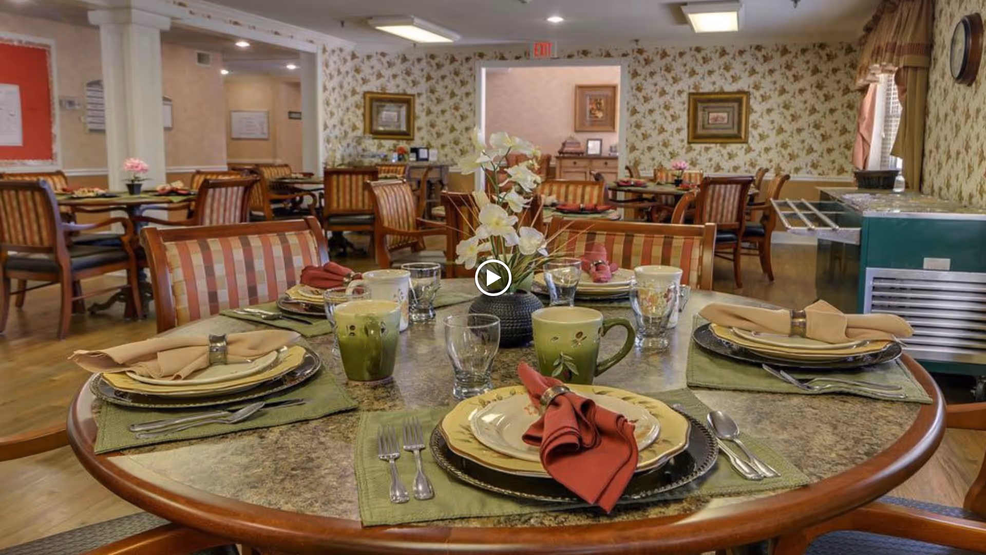 Round dining table set with plates, napkins, mugs, and a floral centerpiece in a decorated senior living dining room.