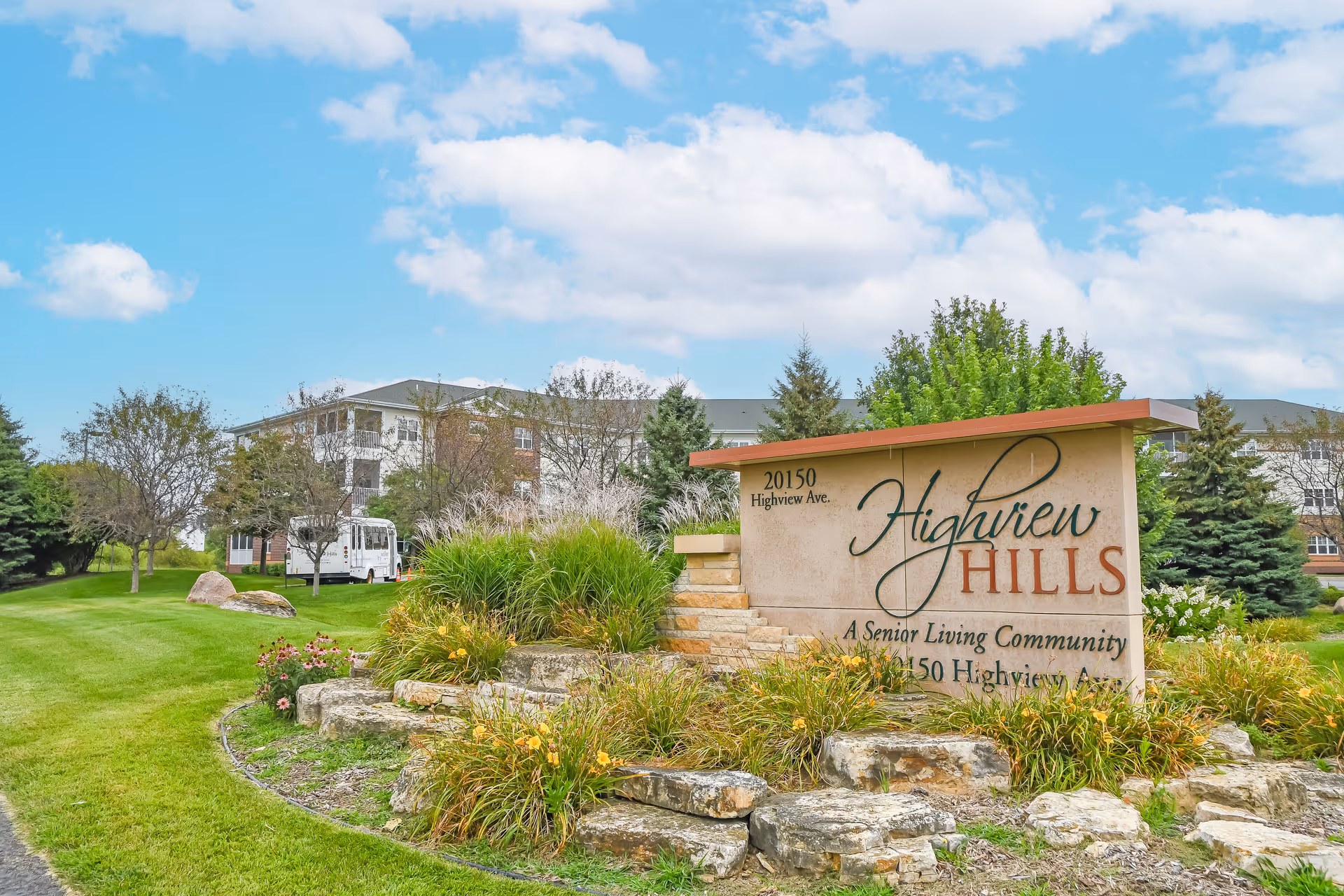Outdoor view of the entrance sign for Highview Hills, a senior living community, surrounded by landscaped greenery, rocks, and trees with a large building and a white shuttle bus in the background under a partly cloudy sky.