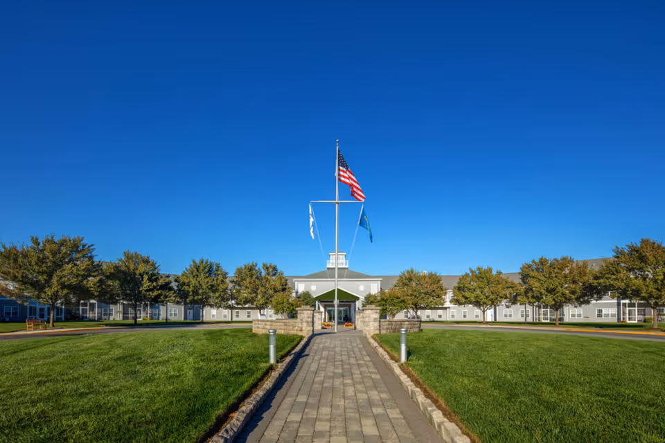 Wide exterior view of The Moorings at Lewes facility with a paved walkway leading to the entrance. The building is surrounded by green lawns and trees under a clear blue sky. Three flags, including the American flag, are flying on flagpoles near the entrance.